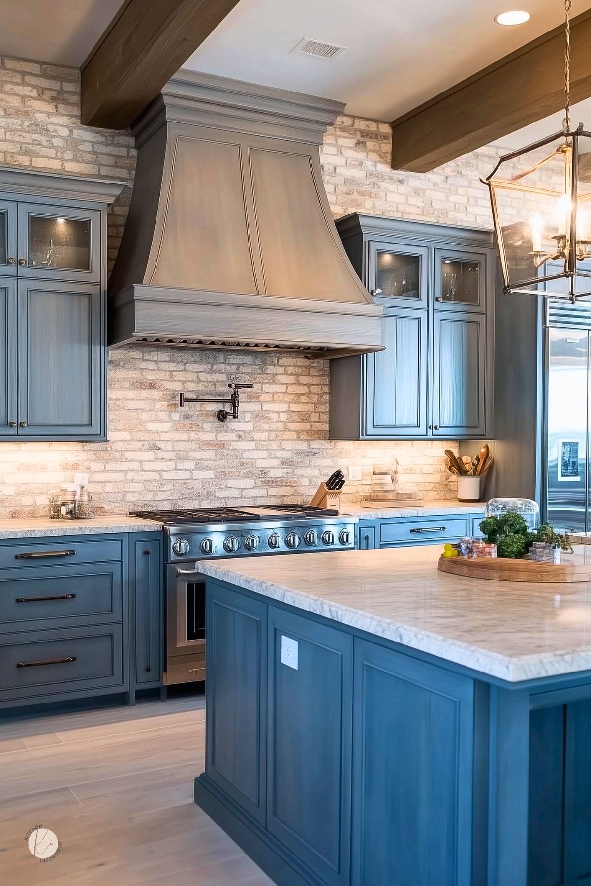 Blue gray kitchen with a whitewashed brick backsplash, matching blue gray cabinets and large island, and a tall taupe wood range hood. Marble countertops, stainless steel range, dark wood ceiling beams, black pot filler, and a lantern pendant light create a polished transitional kitchen design.