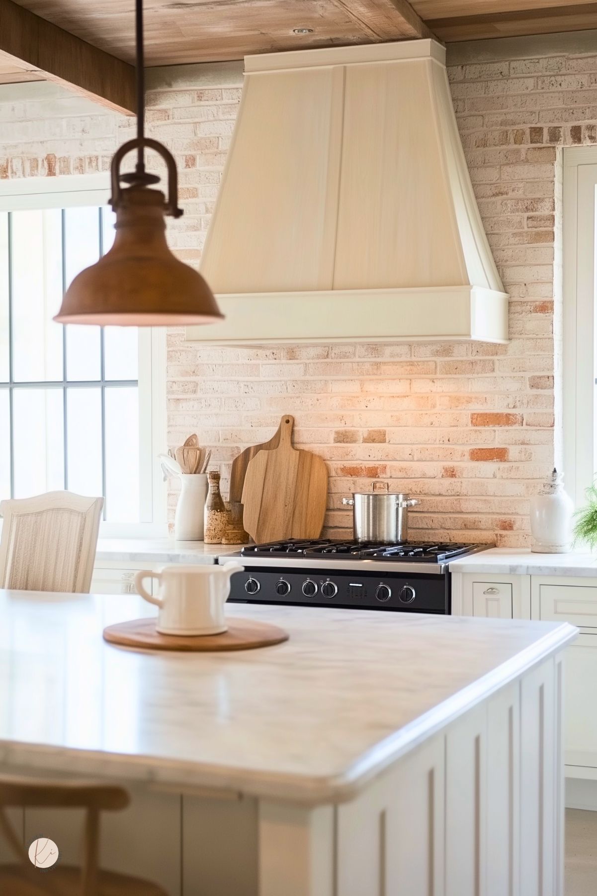 White kitchen with a light painted brick backsplash, pale wood range hood, and white island with a marble-look countertop. Stainless steel range sits below wood cutting boards, while a rustic brown pendant light, soft neutral tones, and bright window create a warm farmhouse kitchen design.
