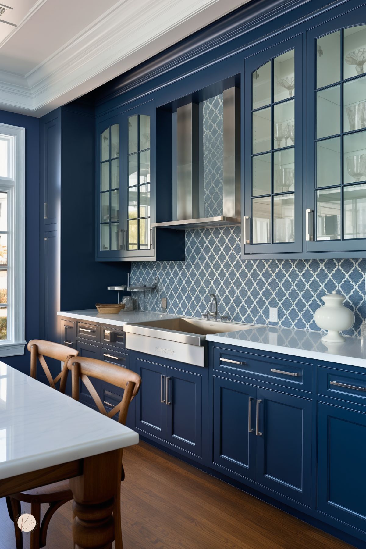 Dark blue kitchen with navy blue glass-front cabinets, white countertops, and a white farmhouse sink. Blue patterned tile backsplash adds texture behind a stainless range hood. Warm wood floors and wood dining chairs soften the space, while large windows and white crown molding keep the kitchen bright.