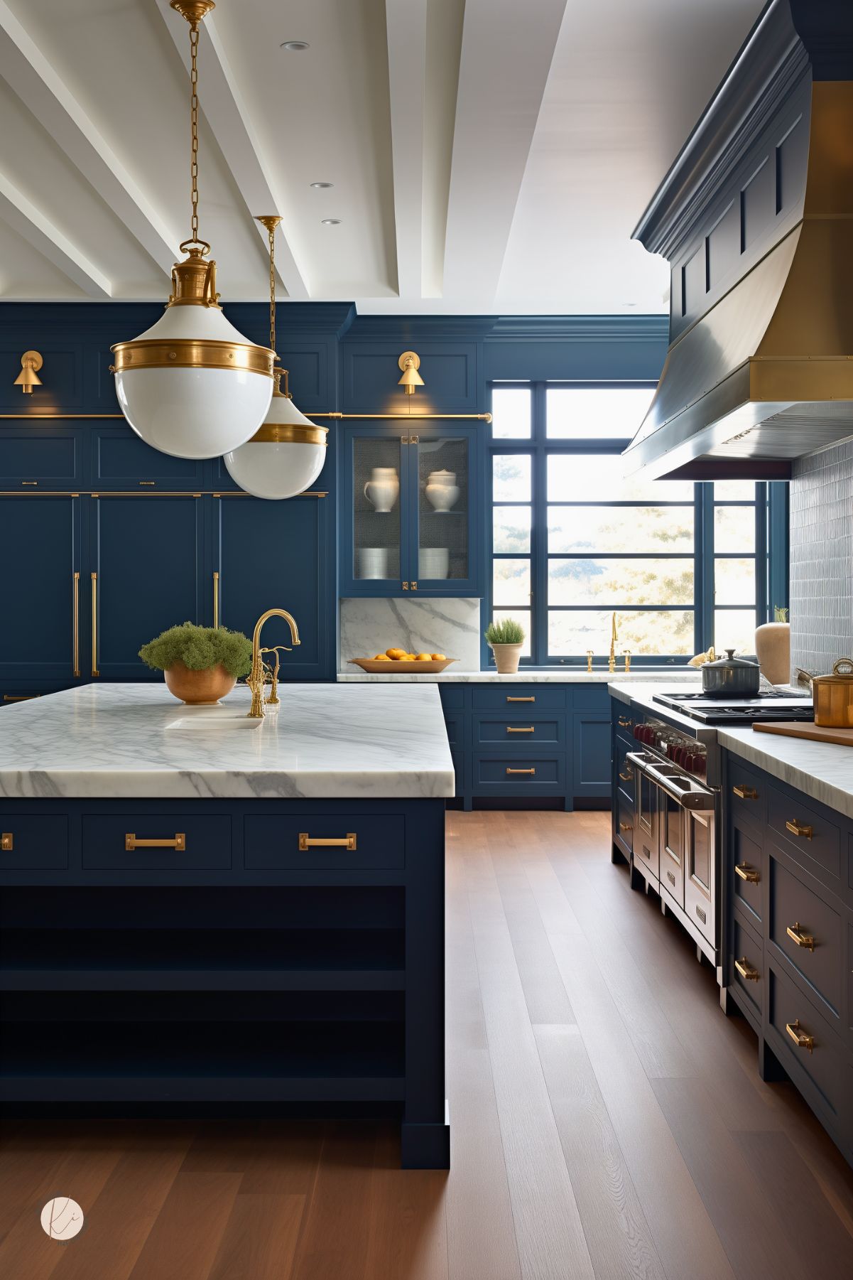 Dark blue kitchen with navy blue cabinets, white marble island countertops, and warm brass hardware. Two large white and brass pendant lights hang over the island, while a matching brass range hood and faucet add a luxe touch. Black-framed windows, light wood floors, and paneled cabinetry give the space a bright, polished look.