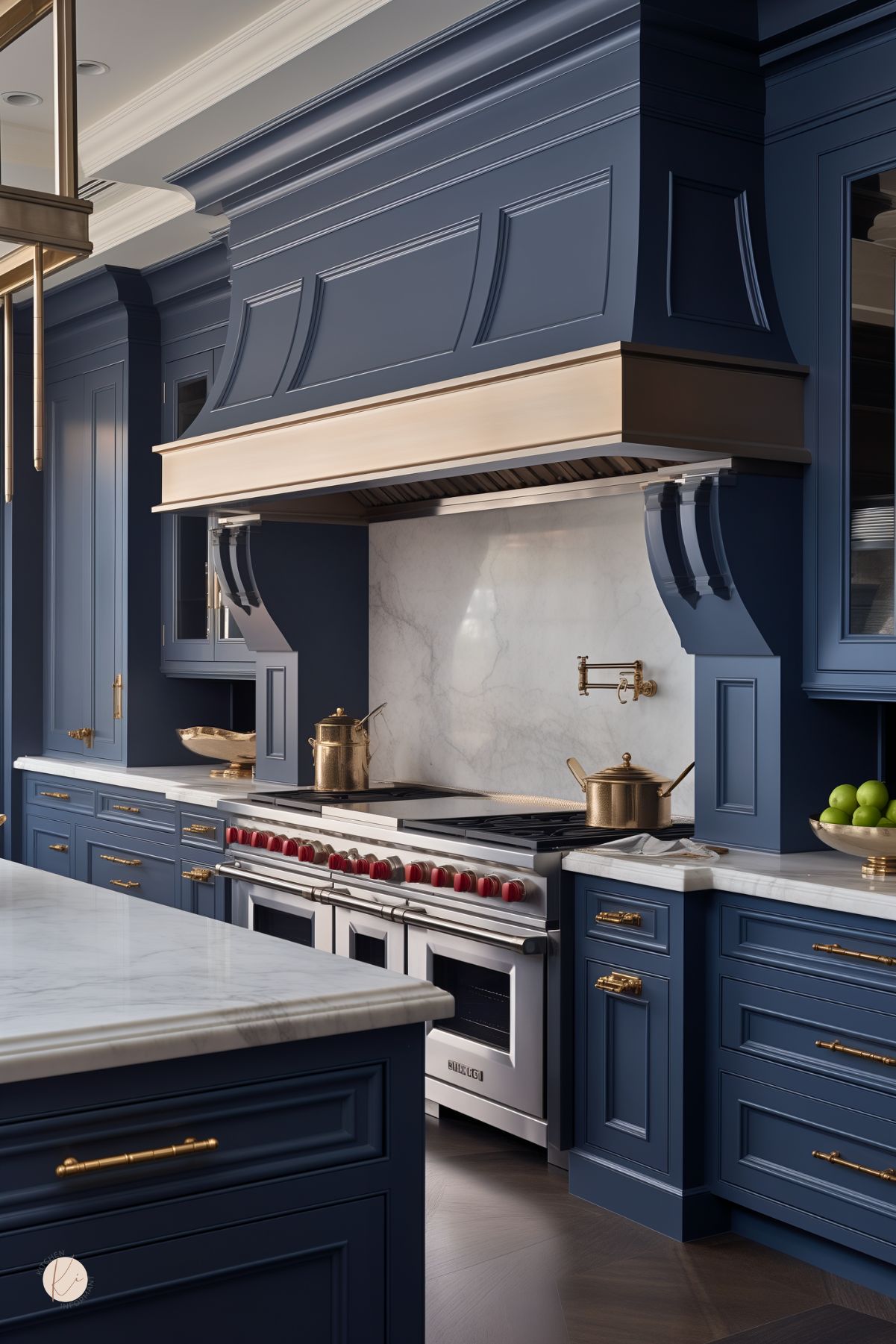 Dark blue luxury kitchen with navy blue paneled cabinets, a dramatic oversized range hood, and white marble countertops and backsplash. A large professional range with red knobs sits below a brass pot filler, with brass cabinet hardware throughout. The marble island and moody blue cabinetry give the kitchen an elegant high-end look.
