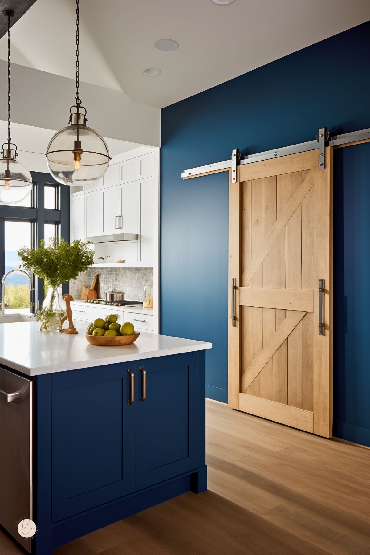 Dark blue kitchen with a navy blue island, white countertops, and a bold blue accent wall. A light wood sliding barn door on black hardware adds contrast. Two glass pendant lights hang above the island, while pale wood floors, a bowl of green apples, and a bright white kitchen in the background keep the space fresh and modern.