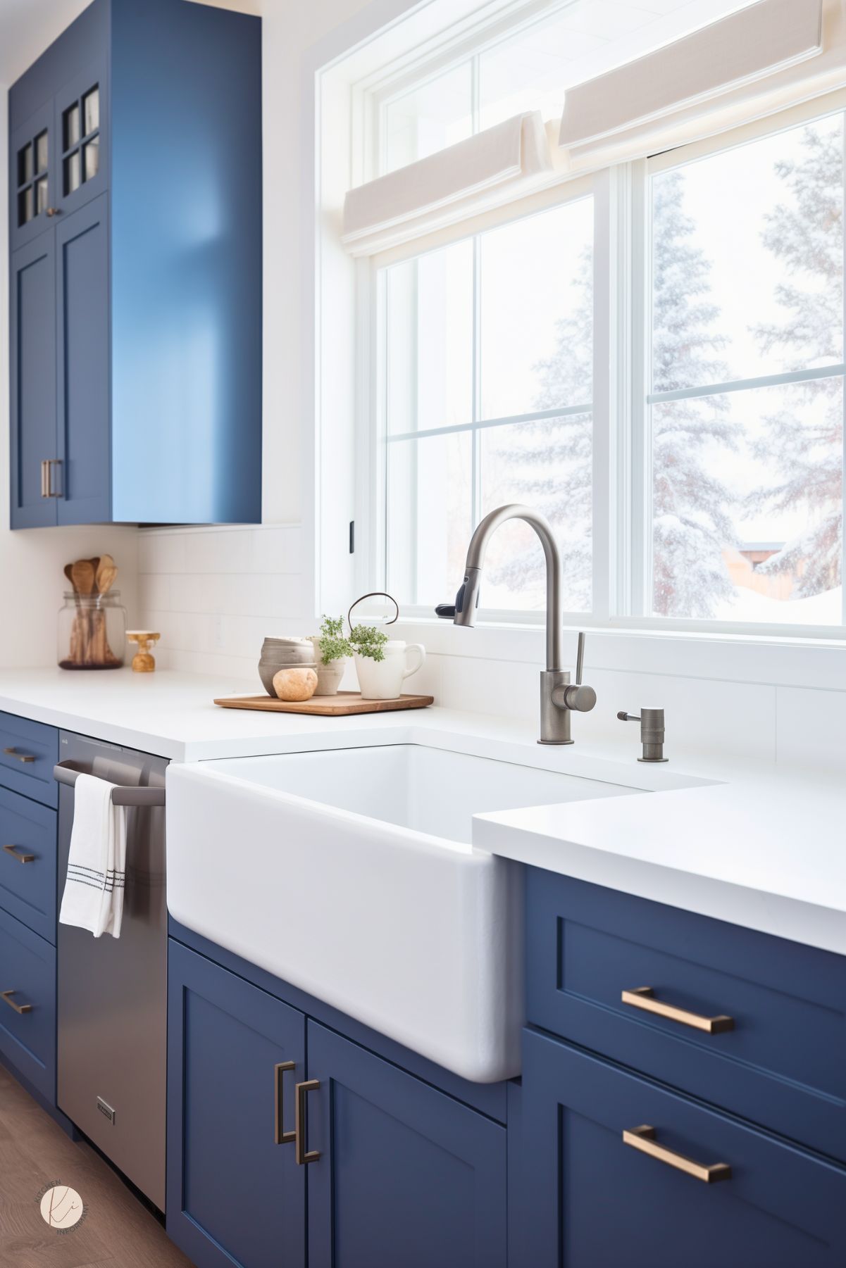 Dark blue kitchen sink area with navy blue shaker cabinets, white countertops, and a large white farmhouse sink beneath an oversized window. Matte nickel faucet, brass cabinet pulls, and a stainless dishwasher add a clean modern touch. Bright natural light, pale wood floors, and simple countertop decor keep the space fresh and airy.