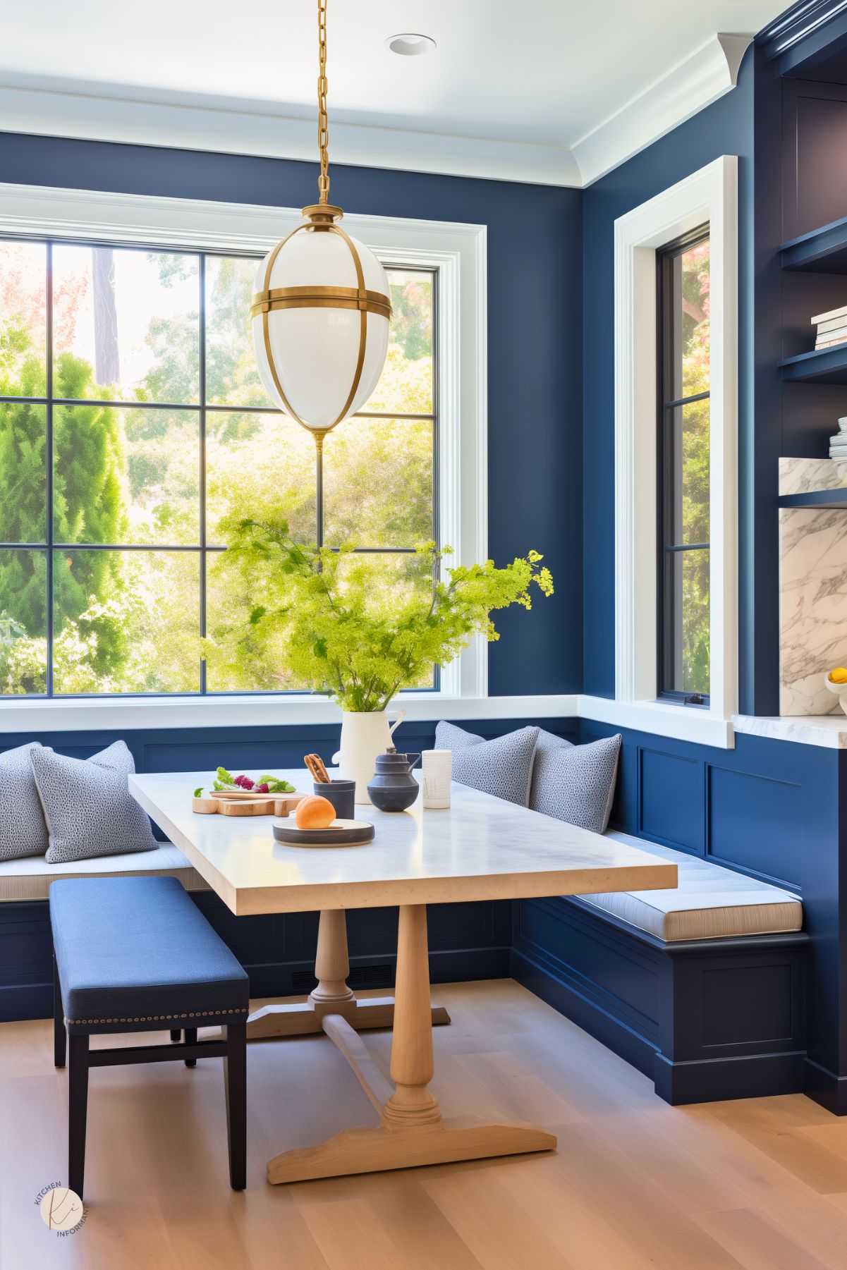 Dark blue kitchen breakfast nook with navy blue paneled walls, built-in banquette seating, and a light wood dining table. Large black-framed windows bring in bright natural light, while a brass and white pendant light adds contrast. Blue cushions, a matching bench, pale wood floors, and simple tabletop decor create a fresh cozy kitchen space.