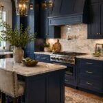 Transitional midnight blue kitchen with tall shaker cabinets, brass hardware, marble countertops, and a marble tile backsplash behind a large matching range hood. Warm lantern pendants, a navy island, stainless steel range, wood floors, and a patterned runner create a polished, upscale kitchen design. Small Kitchen Informant logo at bottom left.