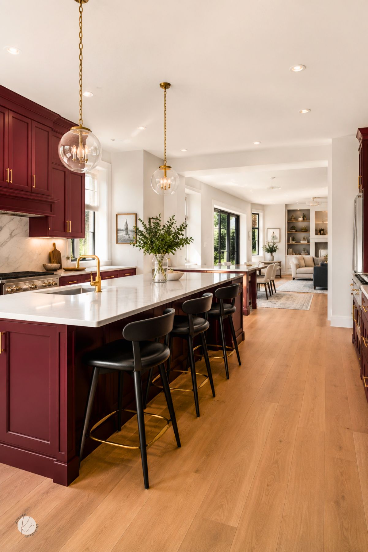 Open concept kitchen with burgundy cabinets, gold hardware, white countertops, black bar stools, glass globe pendant lights, and warm wood floors. A large island leads into a bright dining and living area. Kitchen Informant watermark in the corner.