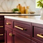 Burgundy kitchen cabinets with gold hardware, white countertops, and a marble backsplash. Close-up of deep red shaker drawers and cabinet doors with brass pulls and knobs, with a small Kitchen Informant watermark in the corner.