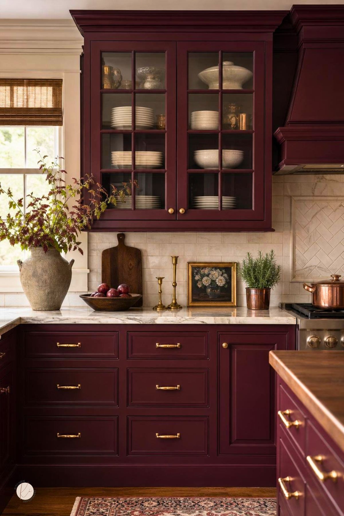 Burgundy kitchen cabinets with gold hardware, glass-front upper cabinets, marble countertops, cream tile backsplash, and a matching range hood. Decorative dishes, copper accents, greenery, and a patterned rug add traditional kitchen style. Kitchen Informant watermark in the corner.