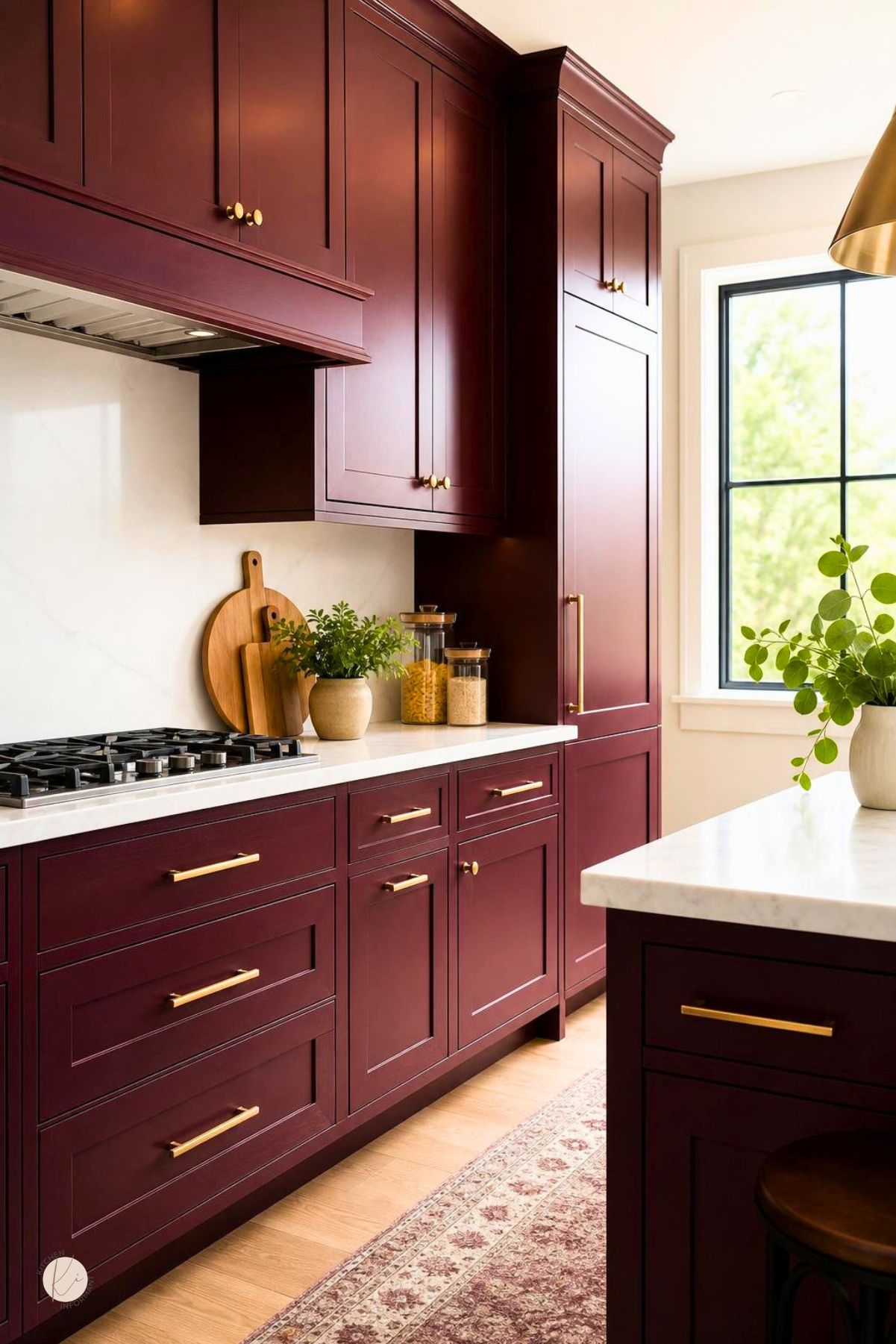 Burgundy kitchen cabinets with gold hardware, white countertops, a matching island, gas cooktop, black-framed window, and warm wood floors. Cutting boards, glass jars, greenery, and a patterned runner add cozy kitchen decor. Kitchen Informant watermark in the corner.