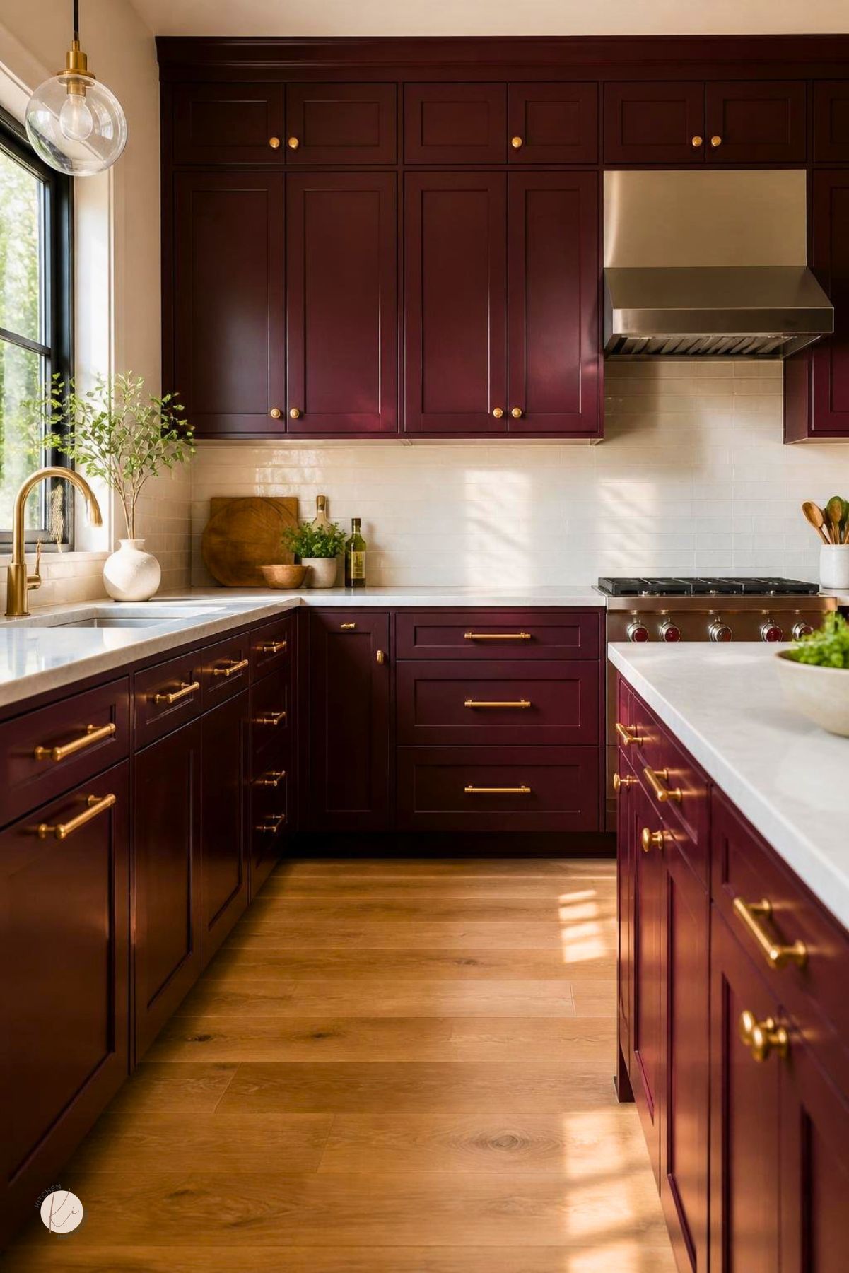 Burgundy kitchen cabinets with gold hardware, white countertops, cream tile backsplash, stainless range hood, brass faucet, and warm wood floors. Sunlight brightens the rich red cabinetry and matching island. Kitchen Informant watermark in the corner.