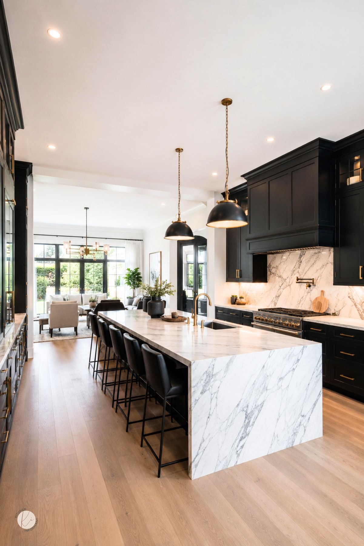 Open kitchen with black cabinets, brass hardware, white marble countertops, and a waterfall island. Black bar stools, brass faucet, black pendant lights, marble backsplash, and light wood floors create a modern luxury kitchen design.