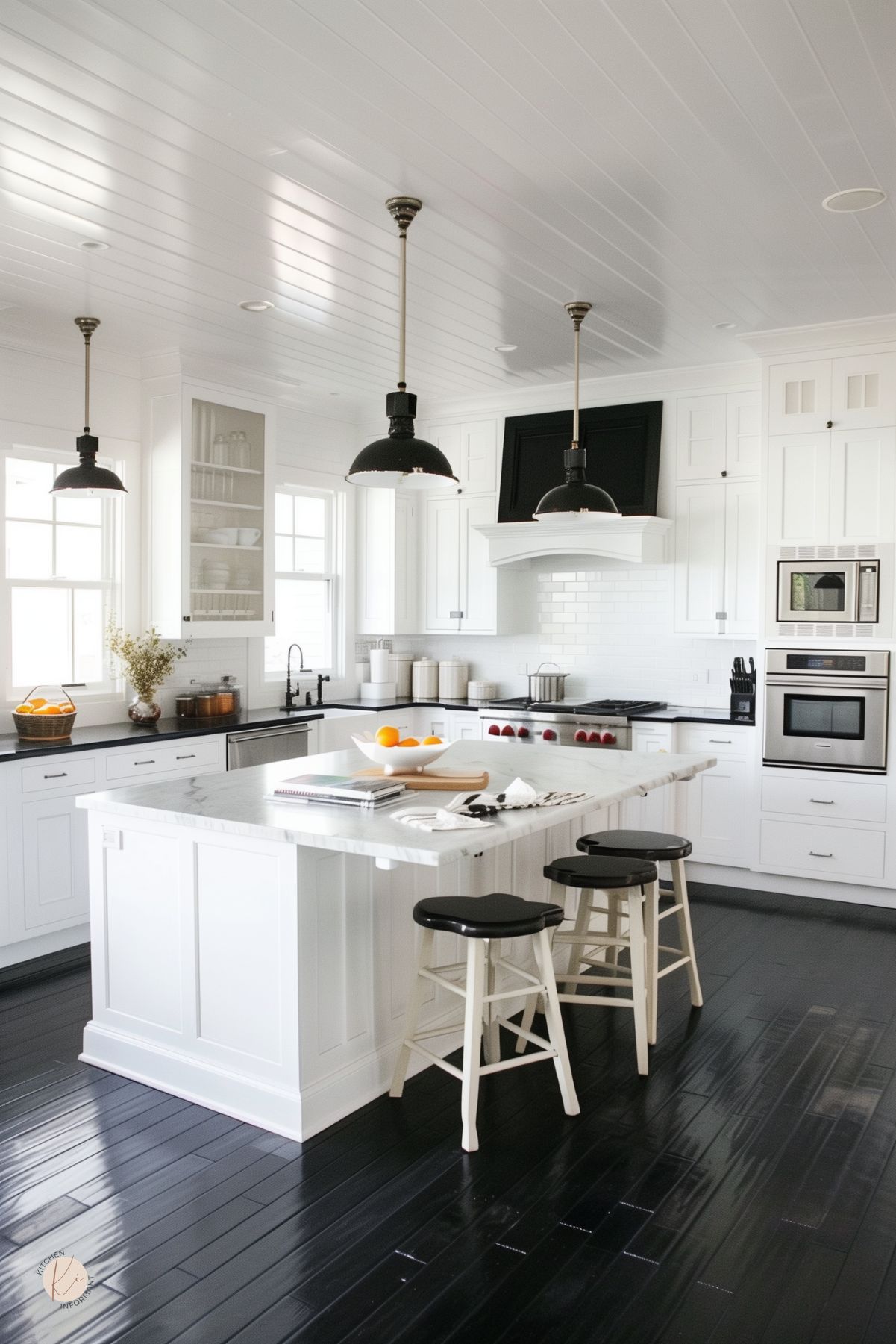 Bright white kitchen with shaker cabinets, marble island, black glossy wood floors, white subway tile backsplash, black pendant lights, black countertops, stainless appliances, white stools with black seats, and a bowl of oranges on the island.