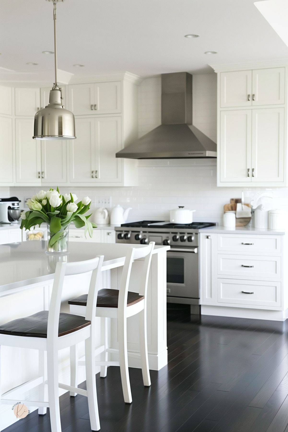 Bright white kitchen with shaker cabinets, glossy black wood floors, stainless range hood, white subway tile backsplash, large island, white bar stools with dark seats, metal pendant light, and white tulips in a glass vase.