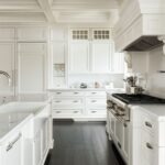 Bright white kitchen with shaker cabinets, coffered ceiling, marble countertops, glossy black wood floors, white paneled range hood, stainless range, glass-front cabinets, large island, chrome hardware, and a clean luxury kitchen design.